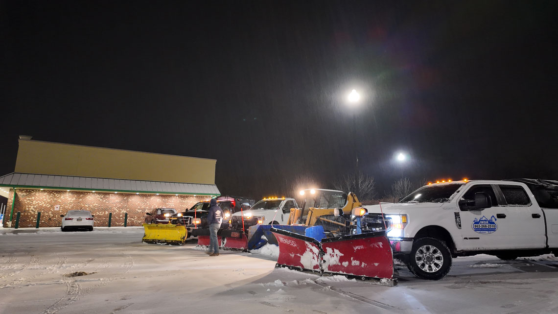 JRW Services plow trucks staged in a commercial parking lot during a night snowstorm